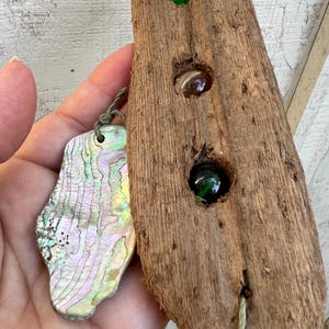 May include: A piece of driftwood with three glass marbles set into it. The marbles are green, brown, and green. A piece of iridescent abalone shell is attached to the driftwood with twine.