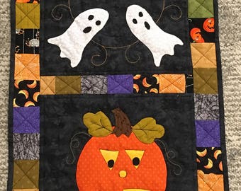 May include: A black quilted table runner with a Halloween theme. It features two orange pumpkins with jack-o'-lantern faces and four white ghosts. The runner is bordered with colorful patchwork squares.