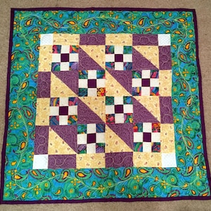 May include: A purple, yellow, and white quilt with a paisley border. The quilt is made up of squares and triangles.