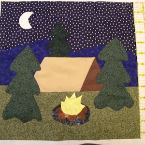 May include: A quilted wall hanging depicting a campsite scene at night. A brown tent with a tan flap is set up between two green trees. A yellow campfire is burning in front of the tent. The sky is dark blue with white stars and a white crescent moon.