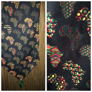 May include: A black table runner featuring a repeating pattern of the African continent in red, yellow, and green geometric designs. The runner has a green tassel at the end and is made of a silky material.
