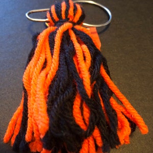May include: A handmade yarn tassel keychain featuring alternating strands of vibrant orange and deep black yarn. The yarn is tightly gathered at the top and secured to a polished silver-tone metal split ring, creating a textured, dangling accessory.
