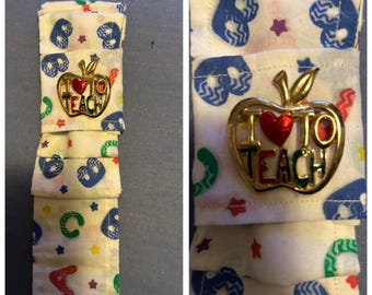 May include: A fabric bookmark with a repeating pattern of blue hearts, green and yellow shapes, and red stars. A gold-colored apple pin with the words "I ❤️ TO TEACH" is attached to the bookmark. The bookmark is made of layered fabric.