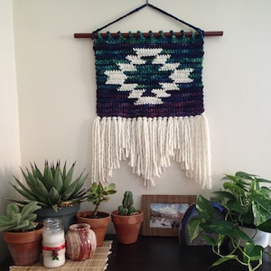 May include: A crocheted wall hanging with a geometric pattern in shades of blue, green, and white. The hanging has a fringe of white yarn and is mounted on a wooden dowel.