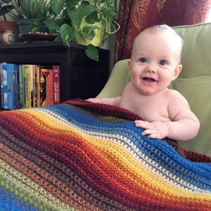 May include: A baby boy with blue eyes and a big smile is sitting in a green chair. He is wrapped in a colorful striped crocheted blanket with shades of red, orange, yellow, green, blue, and white.