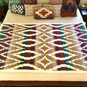 May include: A white crocheted throw blanket with a geometric pattern in shades of brown, green, purple, and gold. The blanket is on a bed with pillows and a wooden headboard.
