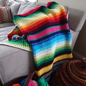 May include: A colorful, handmade crochet blanket draped over a gray sofa. The blanket features horizontal stripes in a rainbow of colors, including red, orange, yellow, green, blue, and purple. A basket of yarn and a woven container are in the foreground.