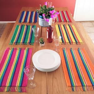 May include: Six colorful crocheted placemats with rainbow stripes and fringe on a wooden table. The placemats are arranged in pairs, with a white plate and wine glass between each pair.