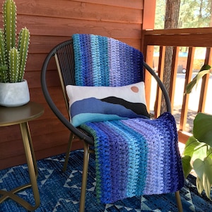 Könnte beinhalten: Ein schwarzer Korbstuhl mit einer blauen und violetten gehäkelten Wolldecke und einem weißen Kissen mit einem Berg- und Sonnenmotiv. Der Stuhl steht auf einer Veranda mit einem Holzgeländer und einer Topfpflanze.