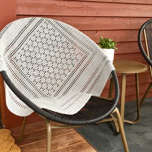May include: A white crocheted throw blanket draped over a black wicker chair with gold metal legs. The chair is on a patio with a wooden floor and a small wooden table.