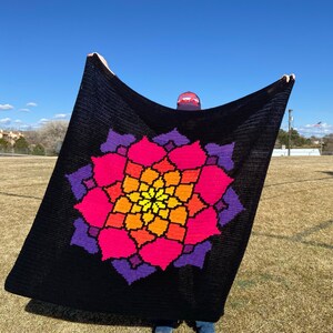 May include: A black crochet blanket featuring a large, colourful mandala design in shades of pink, orange, yellow, and purple. The mandala is centred on the blanket and is surrounded by a black border.