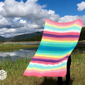 May include: A colorful striped crochet blanket in shades of pink, yellow, green, blue, and purple. The blanket is being held up in front of a lake and mountains.