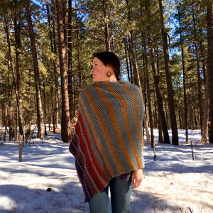 Op de afbeelding: Een vrouw draagt een gestreepte gehaakte poncho in tinten grijs, oranje en paars. De poncho is over haar schouders gedrapeerd en wordt gedragen met een blauwe jeans. De vrouw staat in een besneeuwd bos.