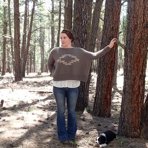 Könnte beinhalten: Eine Frau trägt einen braunen Häkelpullover mit einem geometrischen Muster in einer helleren Farbe. Sie steht in einem Wald mit Bäumen hinter ihr. Ein kleiner schwarz-weißer Hund sitzt im Vordergrund.