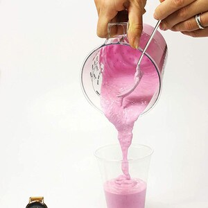 May include: A person pouring pink smoothie from a clear measuring cup into a clear plastic cup. The smoothie is thick and has a swirl pattern. A gold watch is in the lower left corner of the image.
