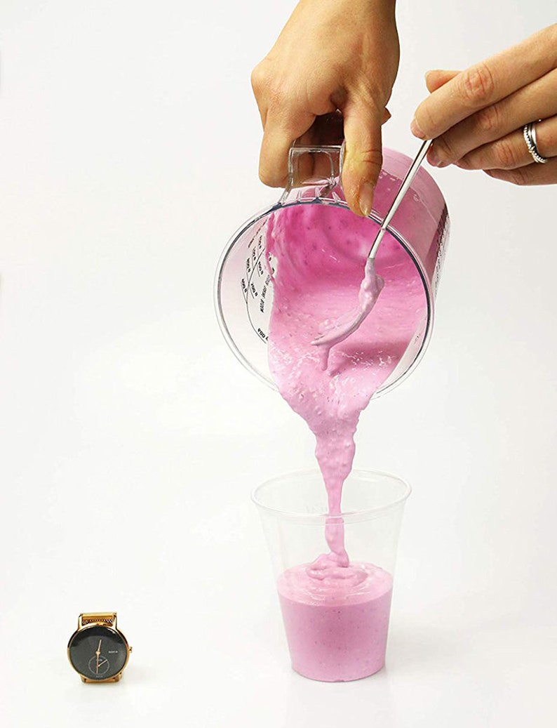 May include: A person pouring pink smoothie from a clear measuring cup into a clear plastic cup. The smoothie is thick and has a swirl pattern. A gold watch is in the lower left corner of the image.