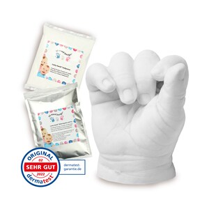 May include: Two white packets of 3D Hand Design Lucky Hand molding powder with a white plaster hand cast of a baby's hand in a fist. The packets have a blue and pink design with a picture of a baby's hand and the text "Lucky Hand Goldpulver". The plaster cast is on a white background. The image also includes a "Dermatest" seal with the text "Original Sehr Gut 2022 Dermatest".