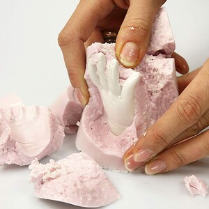 May include: A pink silicone mold is being broken apart to reveal a white ceramic hand sculpture. The mold is textured and has a rough surface.