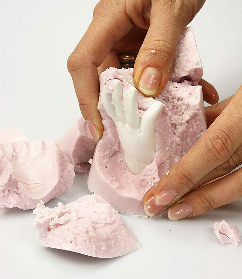 May include: A pink silicone mold is being broken apart to reveal a white ceramic hand sculpture. The mold is textured and has a rough surface.