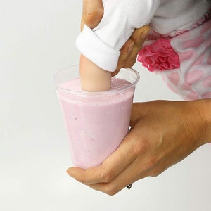 May include: A hand reaches into a clear plastic cup filled with pink liquid. The liquid is thick and creamy, and the hand is pushing down on the surface of the liquid.