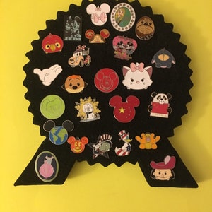 May include: A black pin board shaped like a ribbon with a circle in the center. The board is filled with various Disney character pins, including Mickey Mouse, Minnie Mouse, and other popular characters.