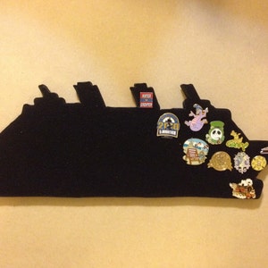 May include: A black felt ship silhouette with various pins attached. The pins feature cartoon characters, logos, and text. The text on the pins includes "25th Anniversary" and "Run Disney".