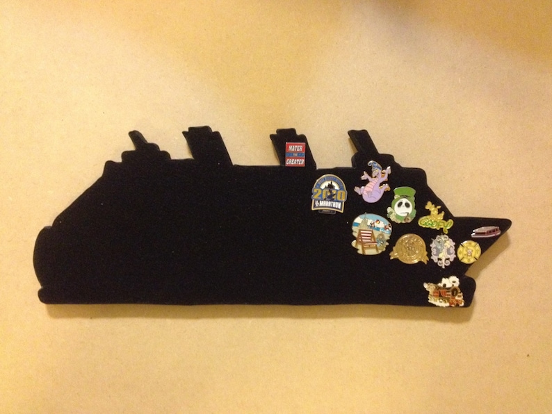 May include: A black felt ship silhouette with various pins attached. The pins feature cartoon characters, logos, and text. The text on the pins includes "25th Anniversary" and "Run Disney".