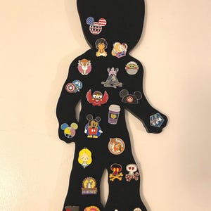 May include: Black silhouette of a person with a variety of colorful pins attached to it. The pins feature characters from popular movies and television shows, including Mickey Mouse, Star Wars, and Disney Princesses.