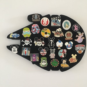 May include: A black Millennium Falcon pin board with a variety of Disney character pins, including Mickey Mouse, Star Wars characters, and other Disney icons.