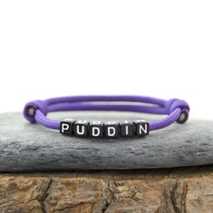 May include: Purple adjustable bracelet with black beads spelling out the word "PUDDIN".