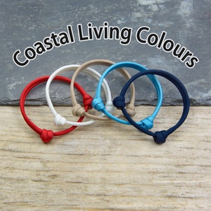 May include: Five colorful bracelets made of braided cord with knotted ends. The bracelets are in red, white, beige, blue, and navy blue. The bracelets are arranged in a circle on a wooden surface. The text "Coastal Living Colours" is written above the bracelets.