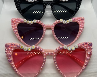 May include: Three pairs of heart-shaped sunglasses with pearl embellishments. The top pair is black with the words "B'DAY GIRL" on the lenses. The middle pair is purple with the words "B'DAY GIRL" on the lenses. The bottom pair is pink with the words "B'DAY GIRL" on the lenses.