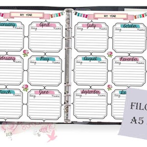 May include: A white planner with a black binder, featuring a monthly calendar layout with spaces for each day and name. The planner is labeled "My Year" and has a decorative border with flags and flowers. The planner is A5 size and is labeled "FILOFAX" on a separate piece of paper.