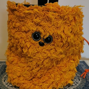 Puede incluir: Una bolsa de magnesio para escalada de color amarillo brillante y peluda con una linda cara de dibujos animados. La bolsa tiene correas negras y un cordón de ajuste negro.