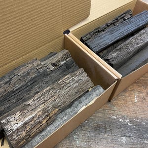 Puede incluir: Dos cajas de cartón abiertas llenas de trozos de madera recuperada. La madera es de color marrón oscuro y tiene una textura rugosa y desgastada.