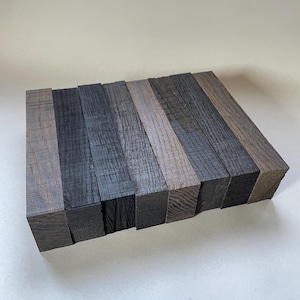 May include: Eight rectangular wooden blocks in shades of grey and black, arranged side by side. The blocks display the natural wood grain and texture. The wood appears freshly cut. Each block is approximately 10 cm in length.