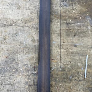 May include: A long, rectangular piece of dark brown wood with a visible vertical grain pattern. The wood features a small, circular marking near the top edge with the number 5 written inside it, indicating a specific piece of lumber.