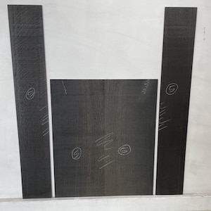 May include: A collection of dark gray wood pieces, including two long, narrow planks and two square pieces. The wood grain is visible, and there are markings in white pencil on the surface. The pieces are arranged against a white background.
