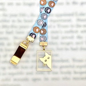 May include: A light blue lanyard with a repeating pattern of paw prints in various colors. The lanyard has a gold-colored metal clasp and a rectangular charm with a cat silhouette.