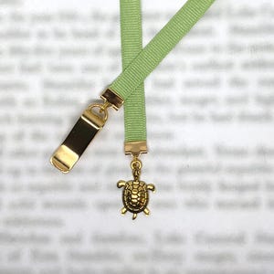 May include: A light green lanyard with a gold-colored metal clasp and a small turtle charm. The lanyard is made of a ribbed fabric and has a rectangular metal piece attached to it.