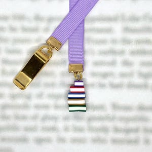 Op de afbeelding: Een lavendelkleurige lintboekmarkering met goudkleurige metalen accenten. De boekenlegger heeft een stapel kleurrijke miniatuurboeken als bedel. Het lint is bevestigd aan een goudkleurige metalen sluiting.