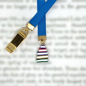 May include: A blue ribbon bookmark with gold-coloured metal accents. The bookmark features a stack of colourful miniature books as a charm. The ribbon is a vibrant blue colour.