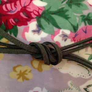 May include: A close-up of a dark green suede cord tied in a knot on a floral fabric background. The fabric has a pink, white, and green floral pattern.
