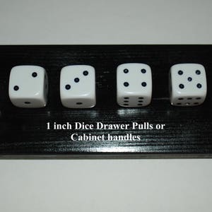 May include: Six white dice drawer pulls or cabinet handles, each with black dots representing numbers from one to six. The dice are 1 inch square and mounted on a black wooden base. Text on the base reads "1 inch Dice Drawer Pulls or Cabinet handles."