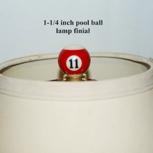 May include: A 1-1/4 inch pool ball lamp finial with the number 11 on it. The ball is red with white accents and is sitting on top of a white lampshade.