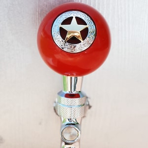 May include: A red beer tap handle with a silver star and decorative design. The handle is attached to a silver metal tap.