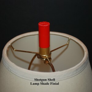 May include: A red shotgun shell lamp shade finial. The finial is attached to a gold metal lamp shade holder.