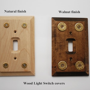 May include: Two wooden light switch covers, one with a natural finish and the other with a walnut finish. Both covers have four brass bullet casings around the switch plate.
