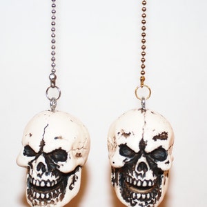 May include: Two white plastic skull-shaped light pull chains with black painted details. The chains are silver and gold.