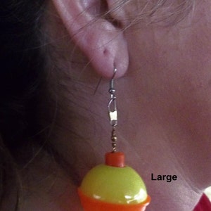 May include: A pair of earrings featuring a yellow and orange bobber charm. The bobber is a popular fishing lure. The earrings are labeled "Large".